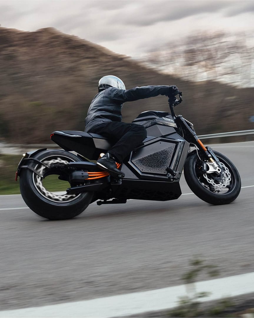 A person wearing a white helmet and black leather jacket rides a modern, black electric motorcycle on a curving road with blurred trees and hills in the background.