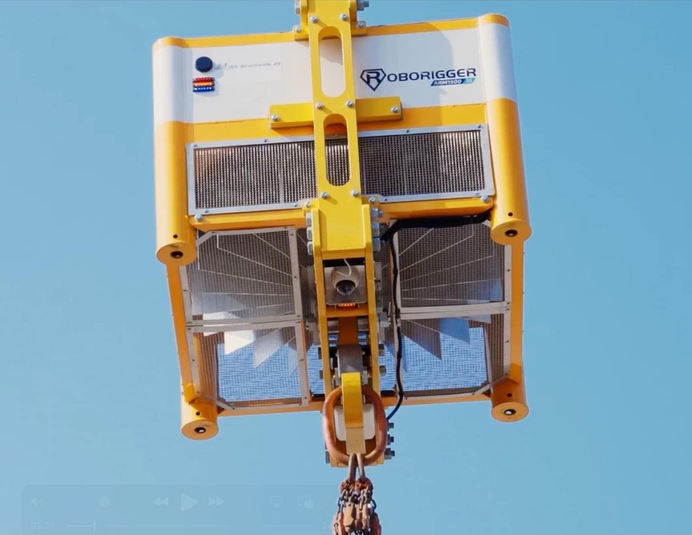 A yellow and white Roborigger device is suspended in the air by a crane hook against a clear blue sky, with visible vents and a control panel on its front.