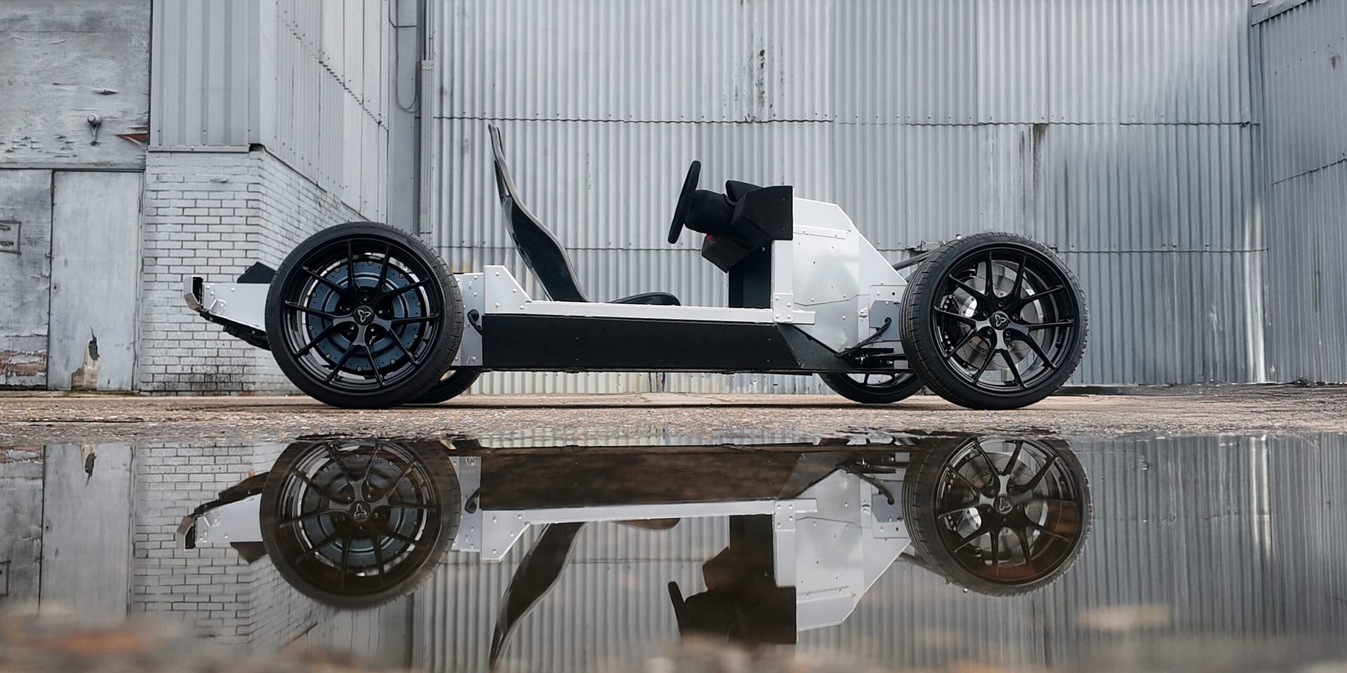 A minimalist, open-frame electric vehicle chassis with large wheels is parked on concrete beside a corrugated metal building, reflected in a nearby puddle of water.